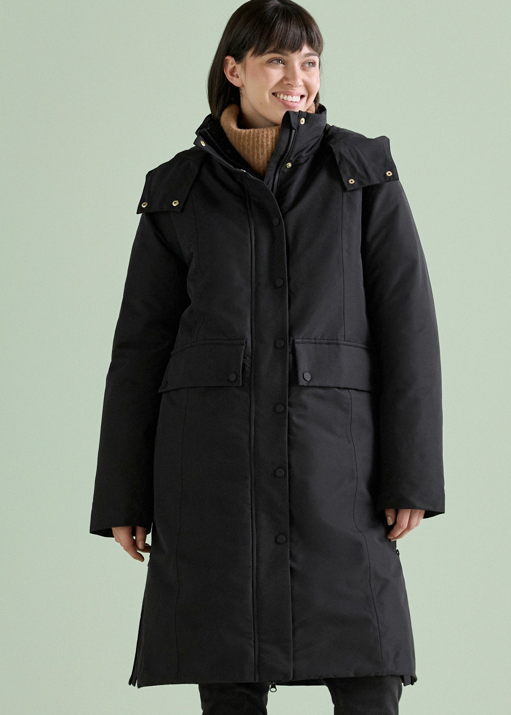 Die Frau trägt einen langen, schwarzen Wintermantel über einem braunen Rollkragenpullover.