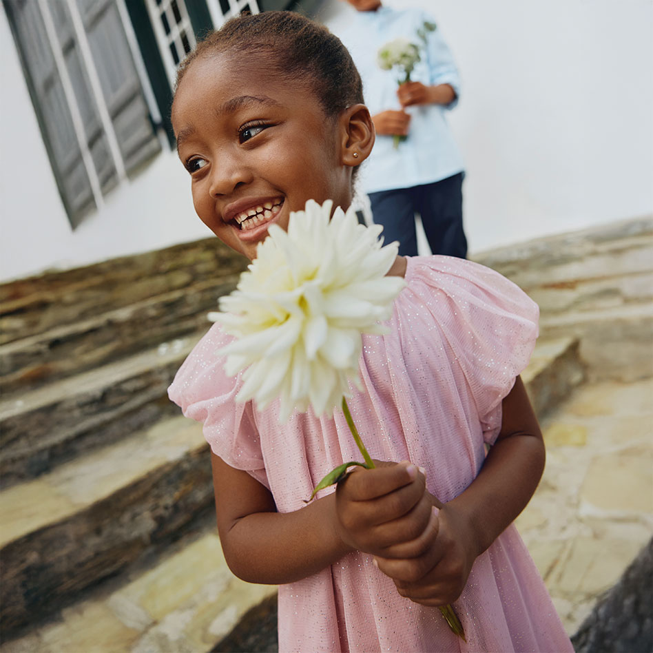 Ein rosa Kleid mit gerüschten Ärmeln schmückt das fröhliche Kind mit der weißen Blume.