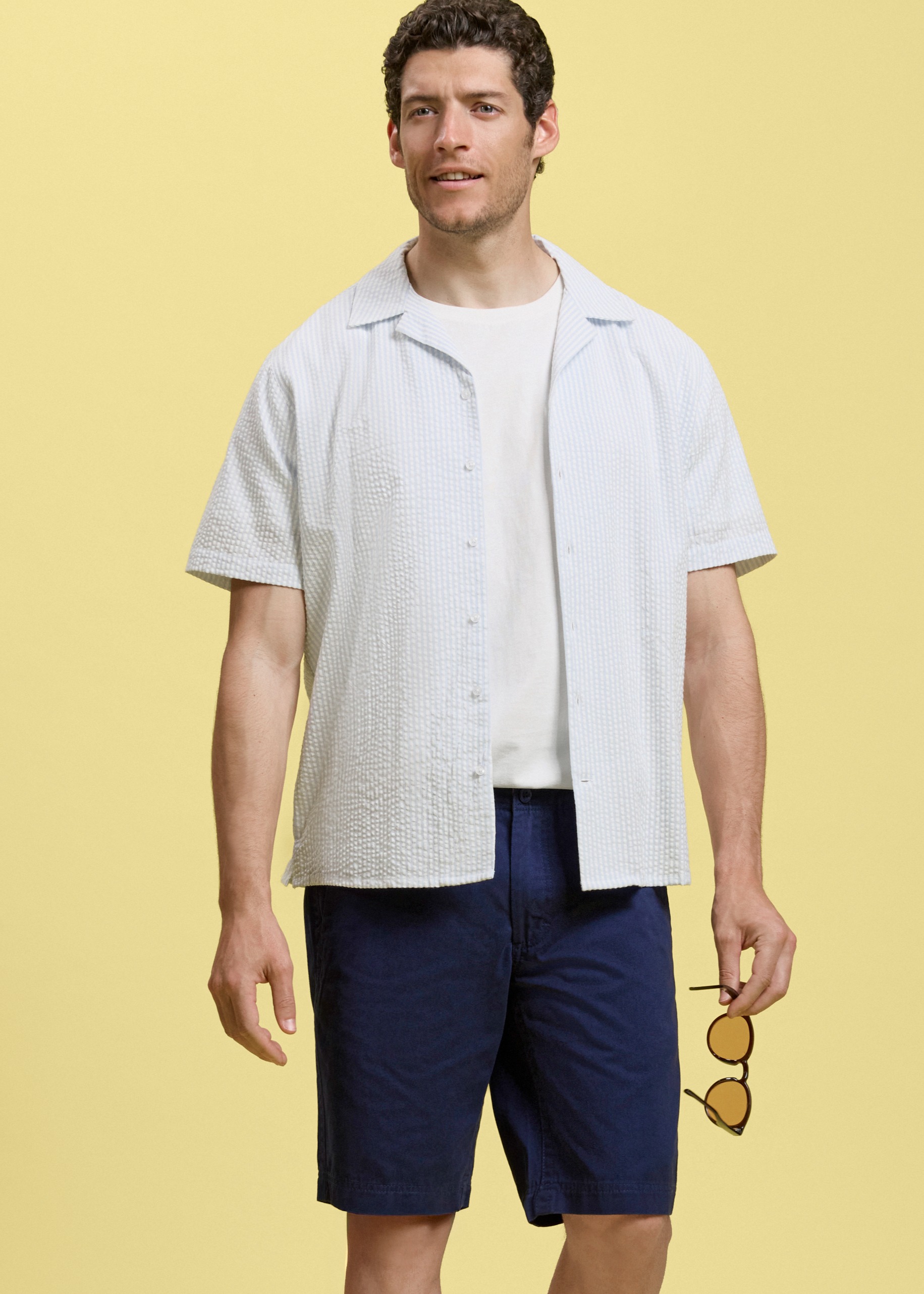 Leichtes, offenes Hemd über weißem T-Shirt, dazu dunkle Shorts und Sonnenbrille.