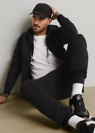 Schwarze Kappe, Hoodie, weißes T-Shirt und schwarze Jogginghose bilden einen lässigen Look.