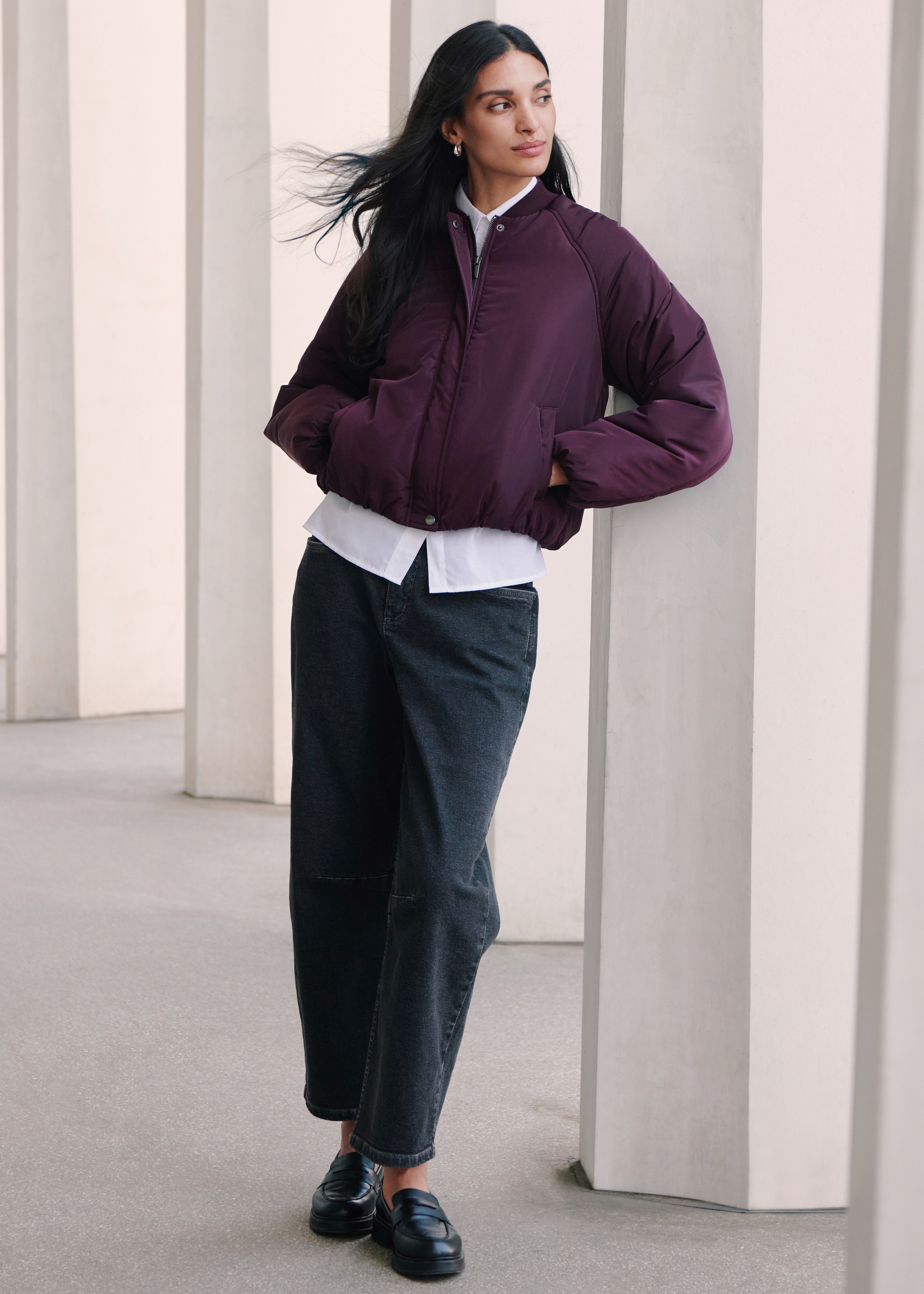 Bordeauxfarbene Steppjacke über weißem Hemd, kombiniert mit dunkler Jeans und schwarzen Slippern.