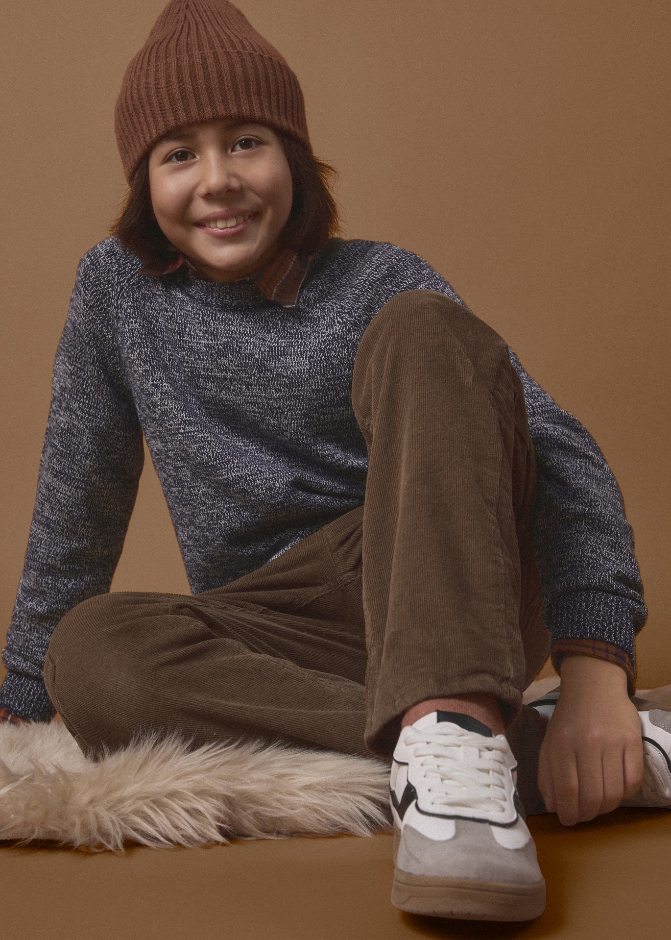 Ein Mädchen trägt eine braune Beanie, einen pinken Pullover, blaue Jeans und weiße Sneaker.
