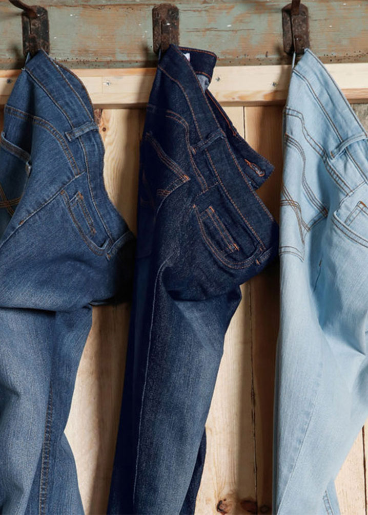 Drei Jeanshosen in verschiedenen Blautönen hängen ordentlich an rustikalen Holzhaken an der Wand.