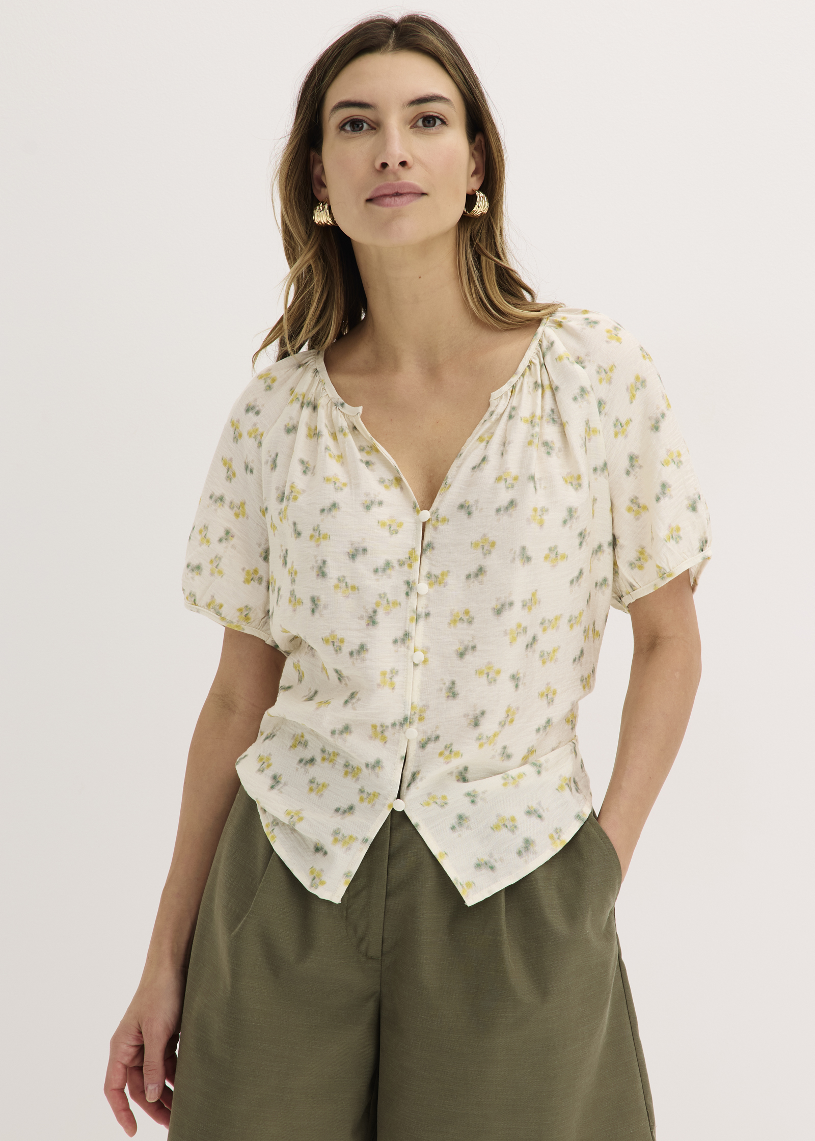 Sie trägt eine helle Bluse mit Puffärmeln und Schmetterlingsmuster, dazu olivgrüne Hosen.