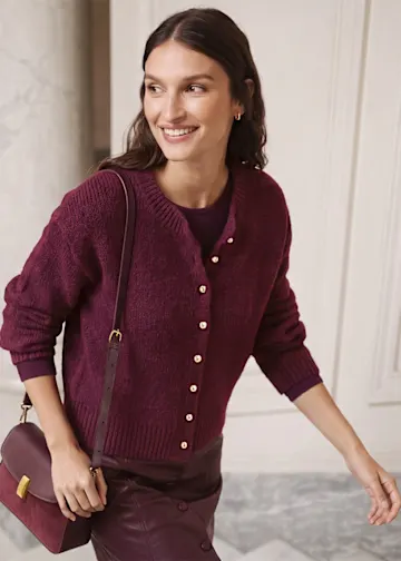 Sie trägt eine bordeauxrote Strickjacke mit Goldknöpfen, dazu passenden Rock und Tasche.