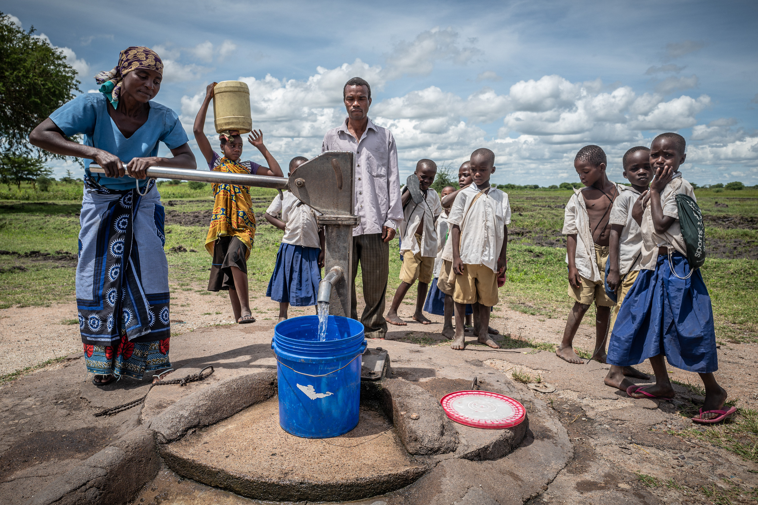 Farbenfrohe traditionelle Kleidung und einfache Schuluniformen prägen das Bild der versammelten Dorfgemeinschaft am Brunnen.