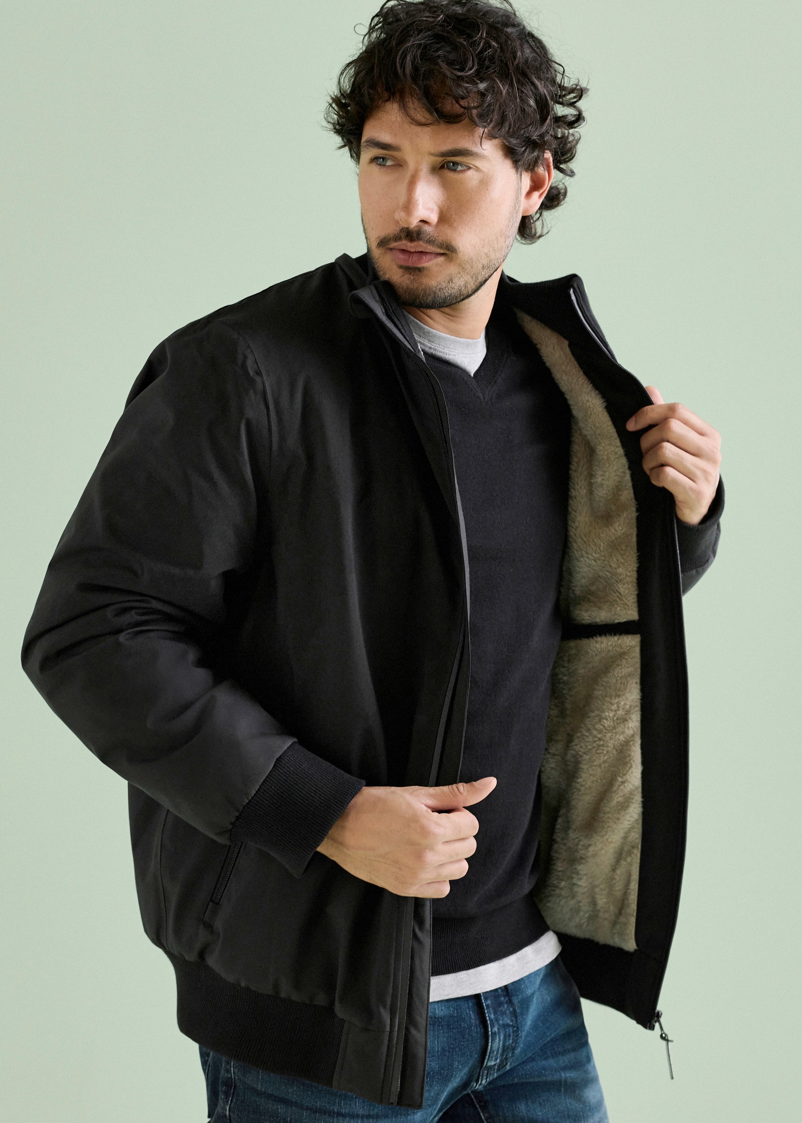 Schwarze Bomberjacke mit flauschigem Futter, dunkler Pullover und Jeans.