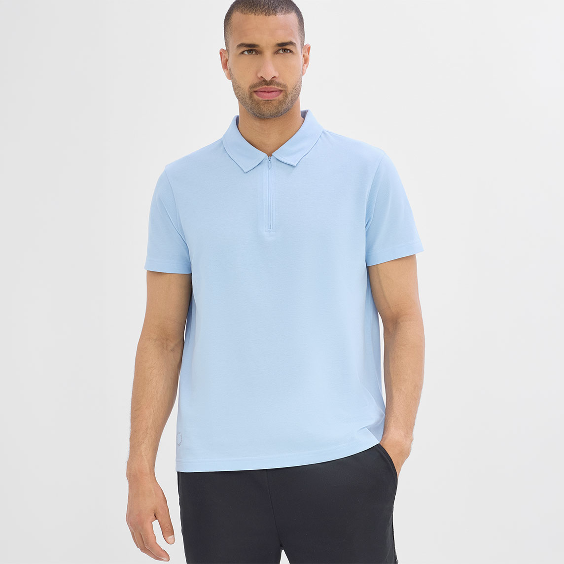 Der Mann trägt ein hellblaues Poloshirt mit Reißverschluss und schwarze Hose.