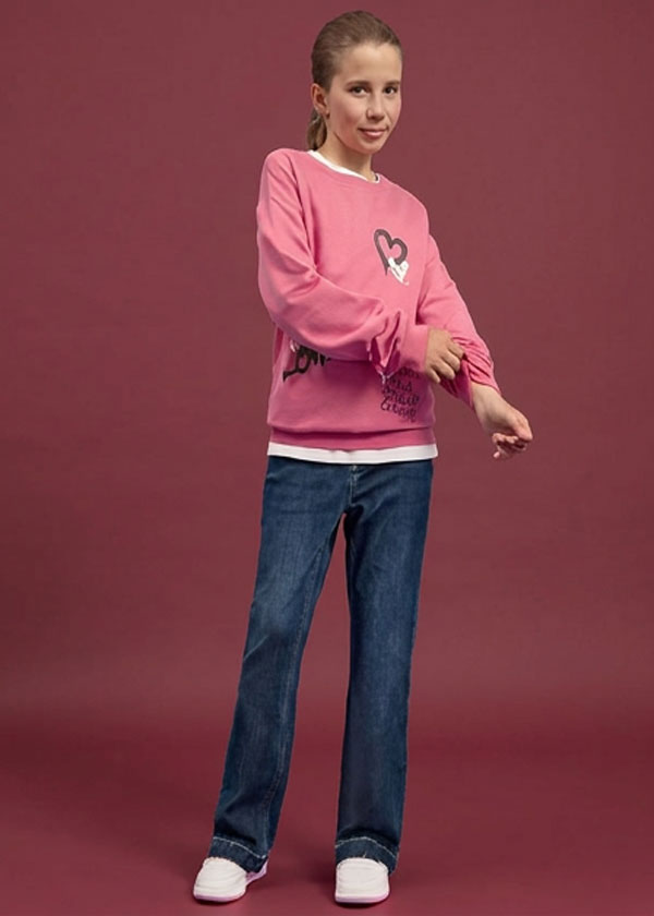 Sie trägt ein pinkes Sweatshirt mit Herzmotiv, blaue Jeans und weiße Sneaker.