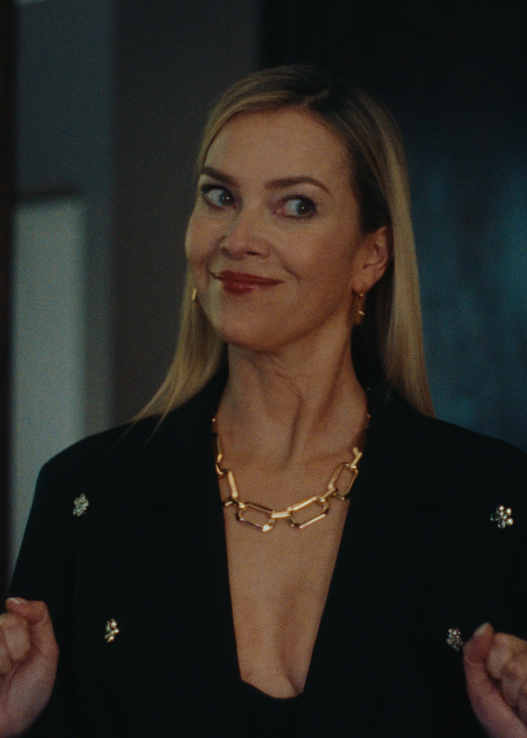 Sie trägt einen eleganten schwarzen Blazer mit funkelnden Details und einer auffälligen Goldkette.