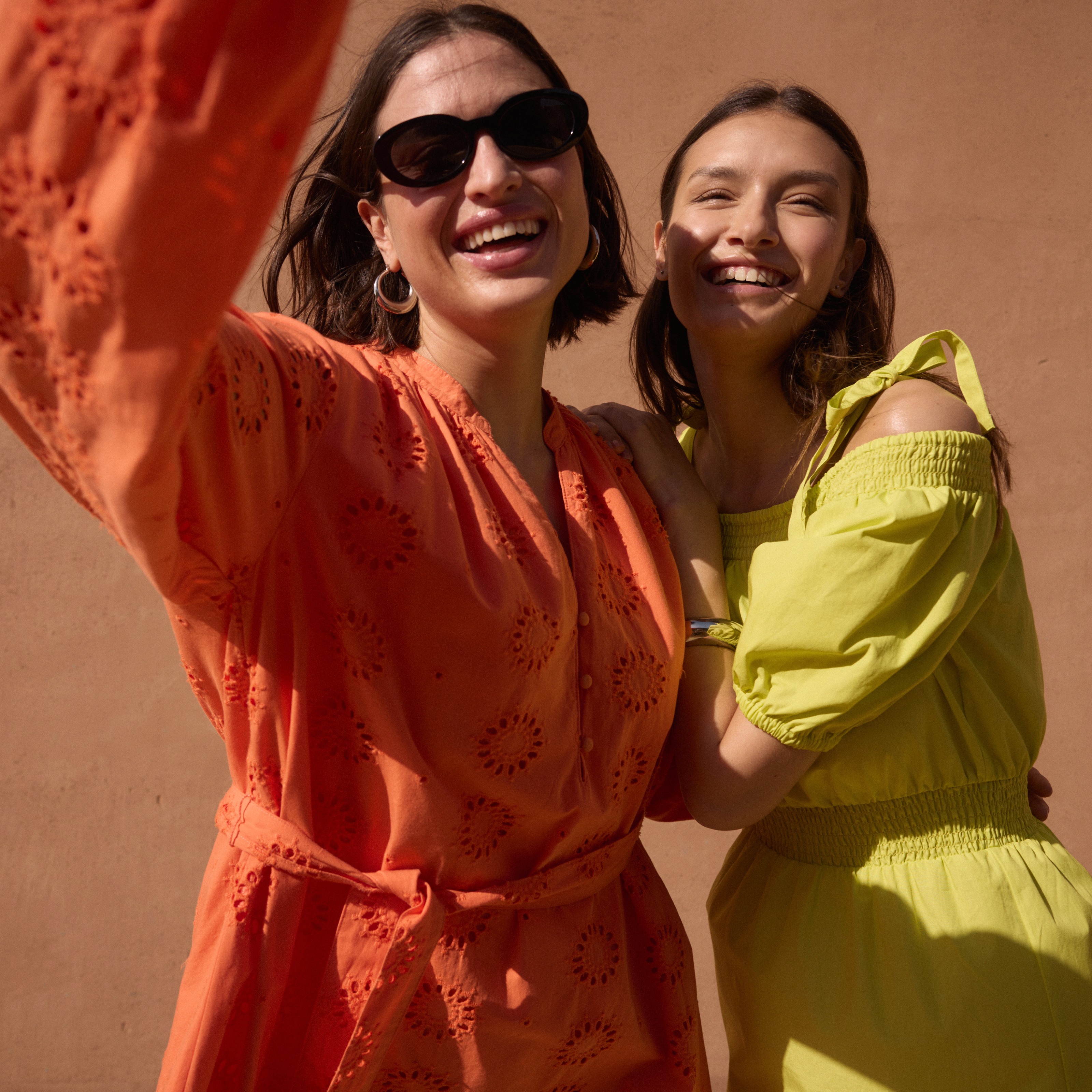Sommerliche Kleider in leuchtendem Orange und Neongelb. Sonnenbrille und breites Lächeln vervollständigen den Look.