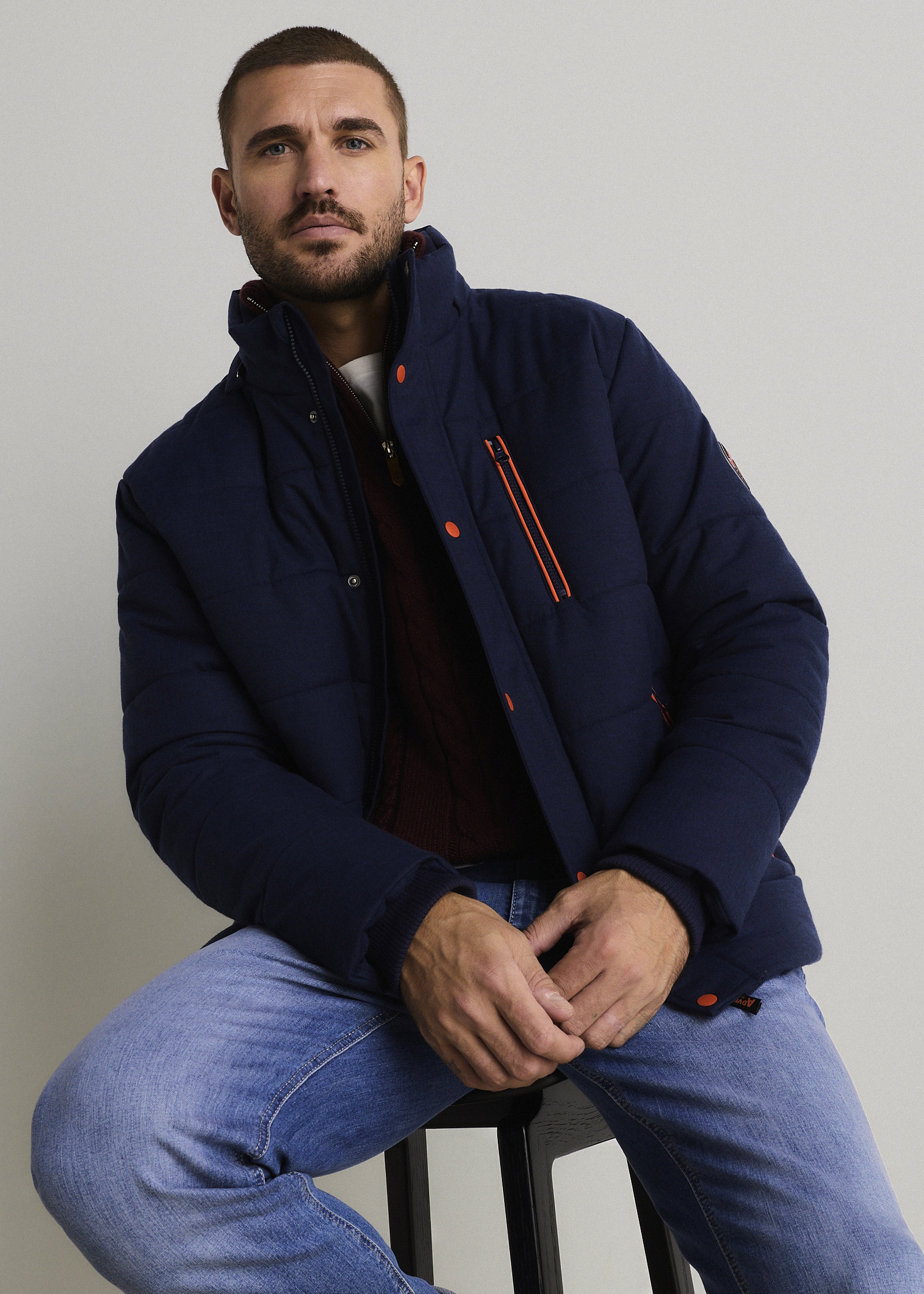 Dunkelblaue Steppjacke mit orangefarbenen Details, roter Strickpullover und helle Jeans.