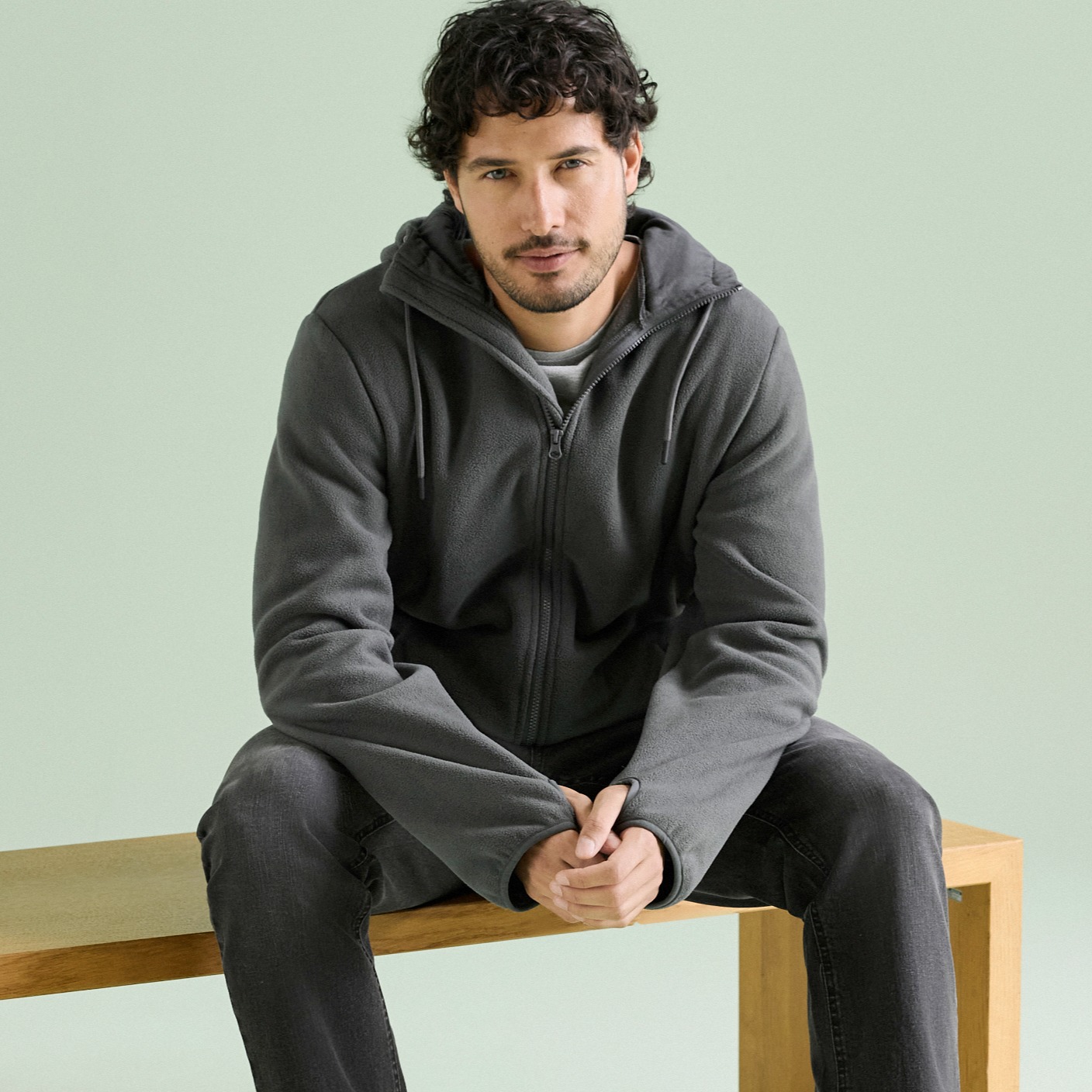 Warmer grauer Kapuzenpullover, helles T-Shirt und dunkle Jeans kennzeichnen sein Outfit.