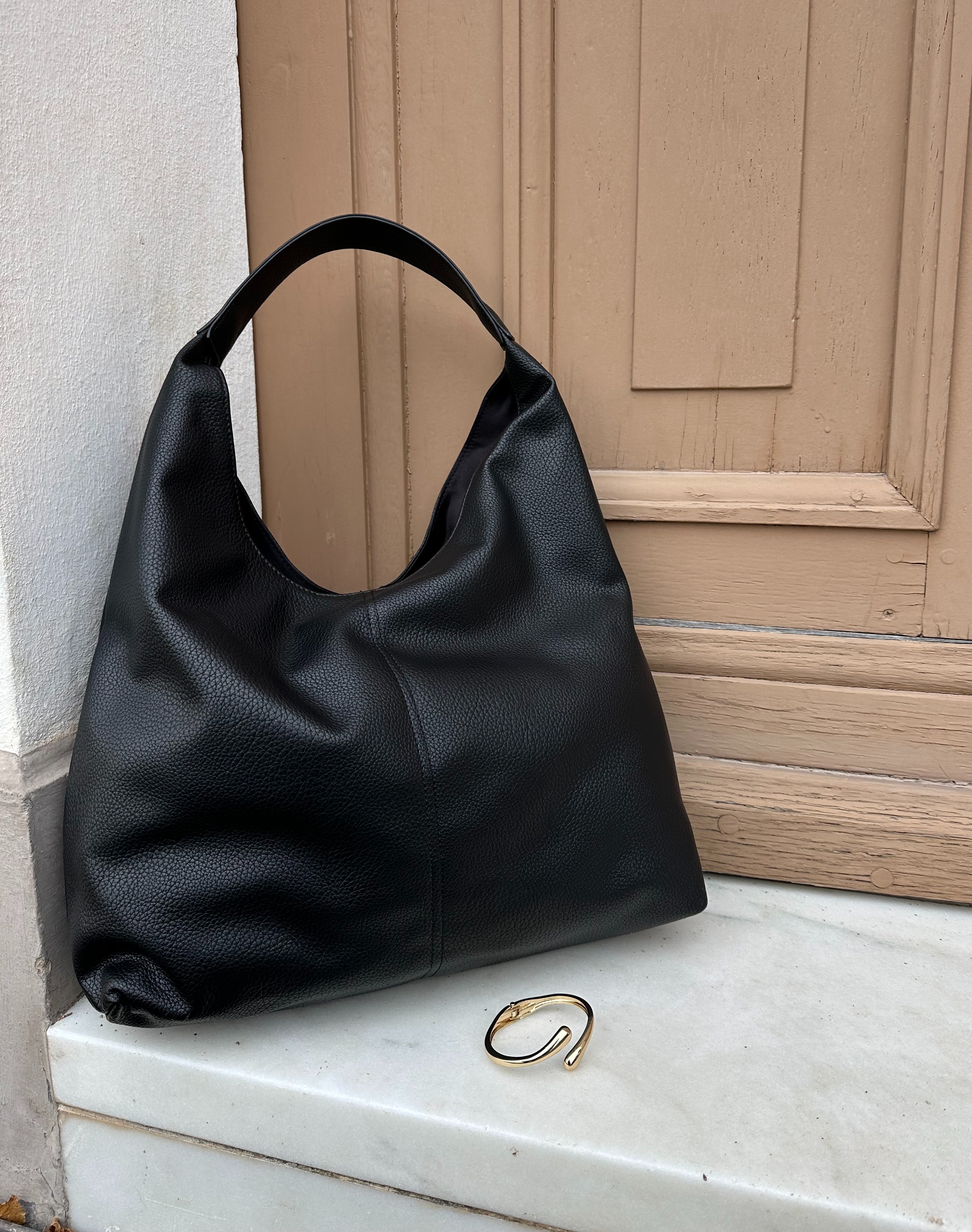 Elegante schwarze Lederhandtasche neben goldenem Armreif auf weißer Türschwelle.