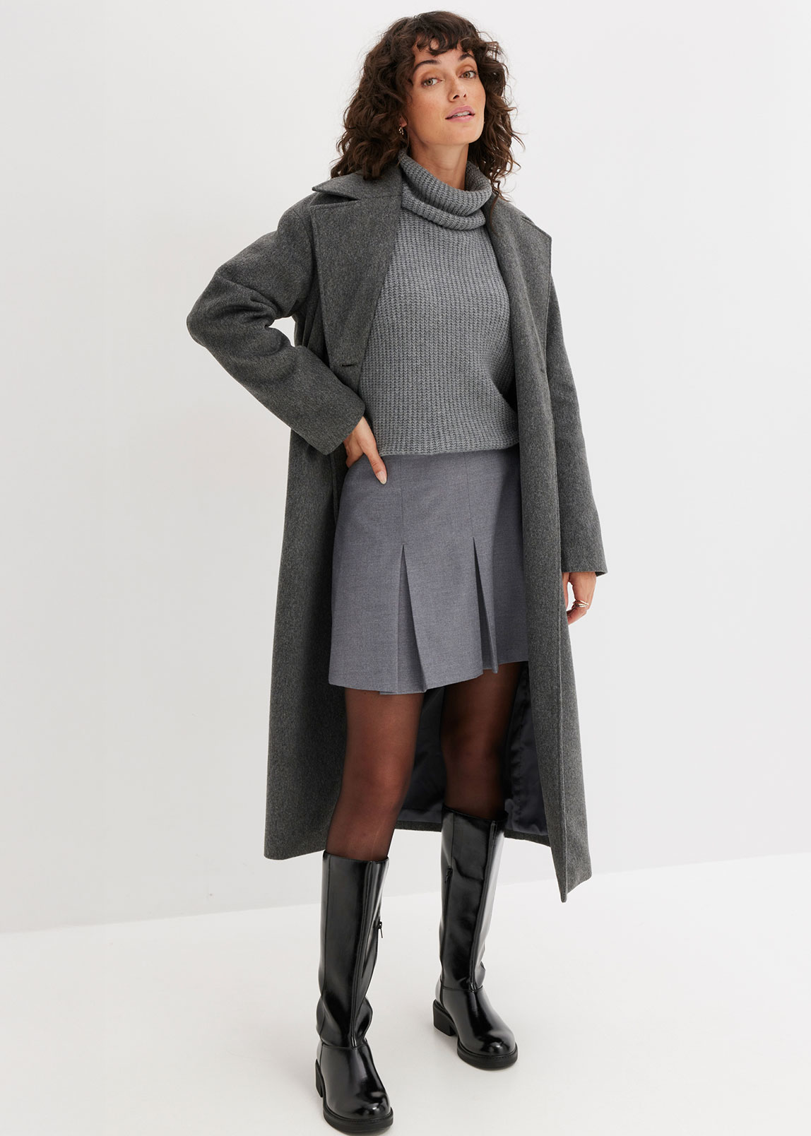 Grauer Wollmantel, Rollkragenpullover, Faltenrock und schwarze Lederstiefel.