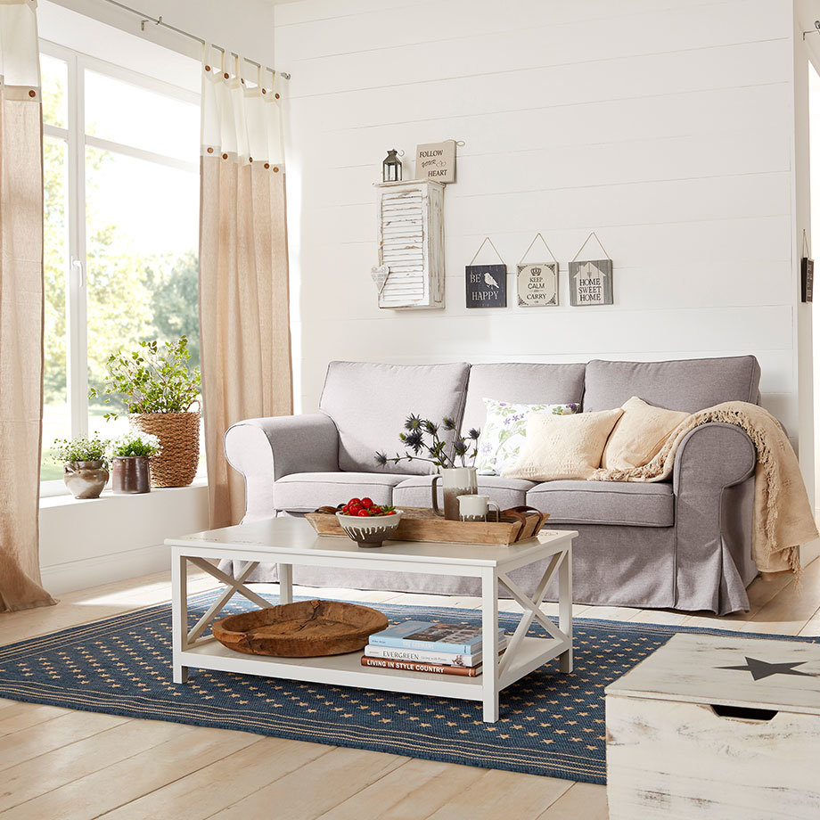 Helles Wohnzimmer mit grauem Sofa, beigen Vorhängen und blauem Teppich. Gemütliche Atmosphäre durch weiche Textilien.