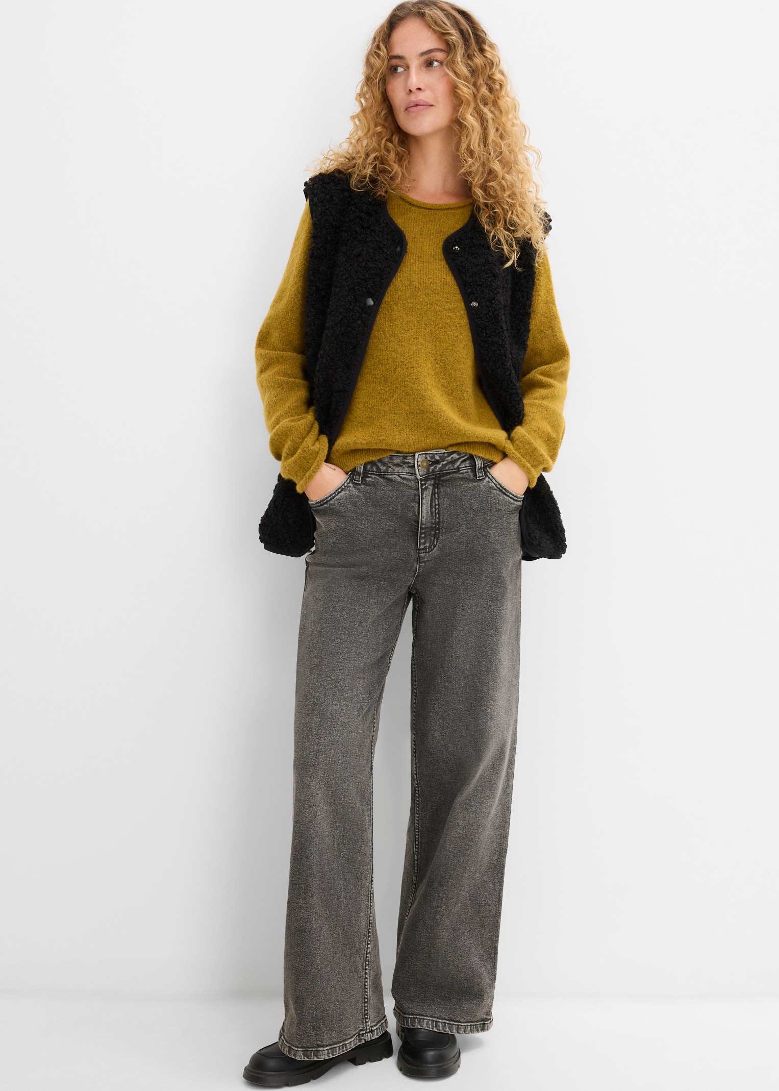 Die Kleidung besteht aus einem senfgelben Pullover, schwarzer Weste und grauen Jeans.