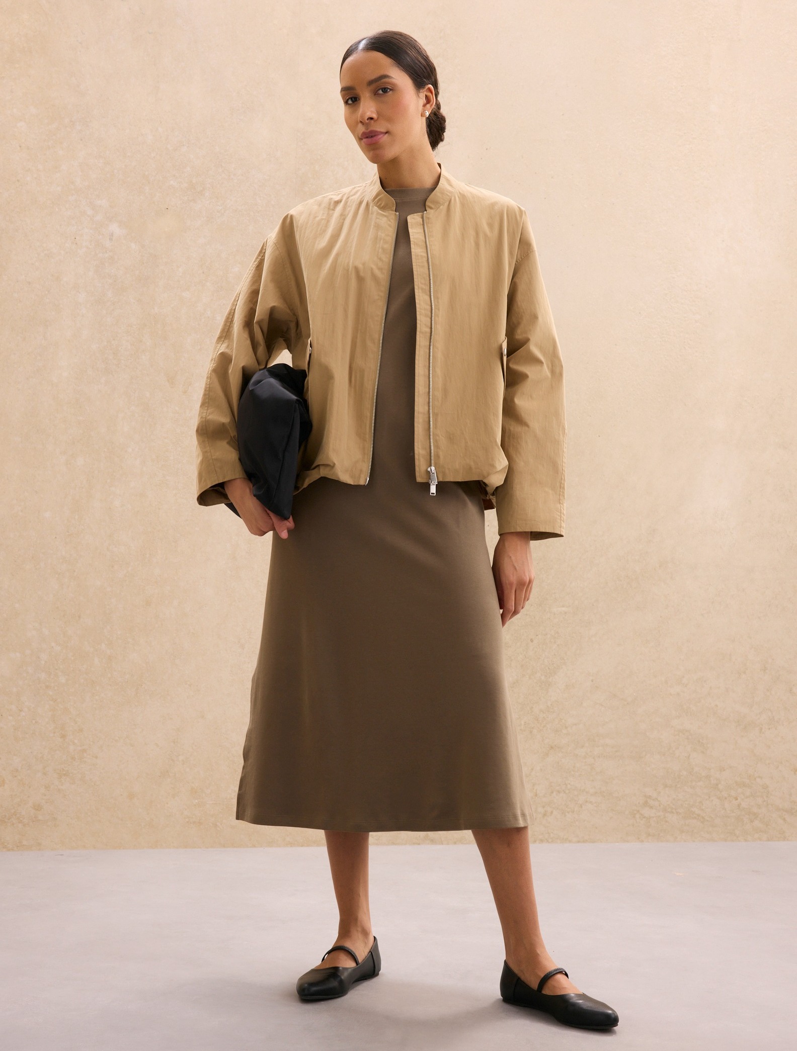 Elegante beige Jacke über braunem Kleid. Schwarze Clutch und flache Schuhe vervollständigen den Look.