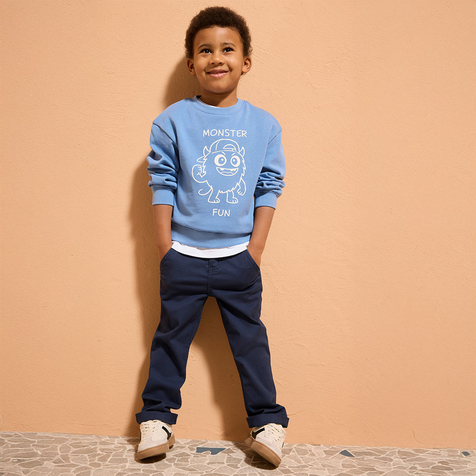 Hellblauer Pullover mit weißem Aufdruck, dunkle Jeans und beige Schuhe bilden ein lässiges Outfit.