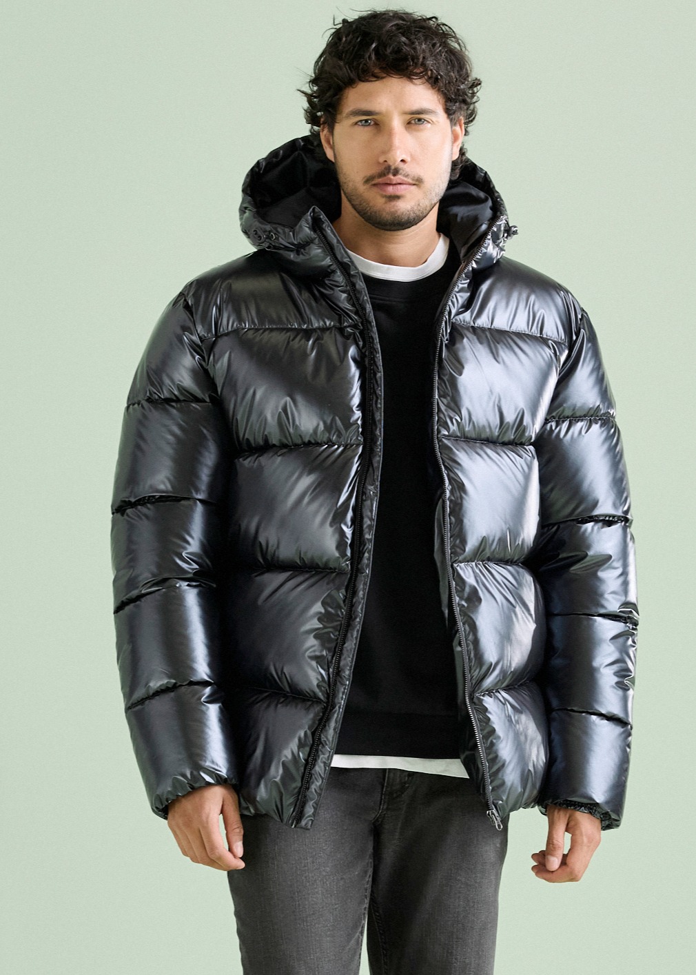 Der Mann trägt eine stylische, glänzende schwarze Pufferjacke mit Kapuze und dunkler Hose.