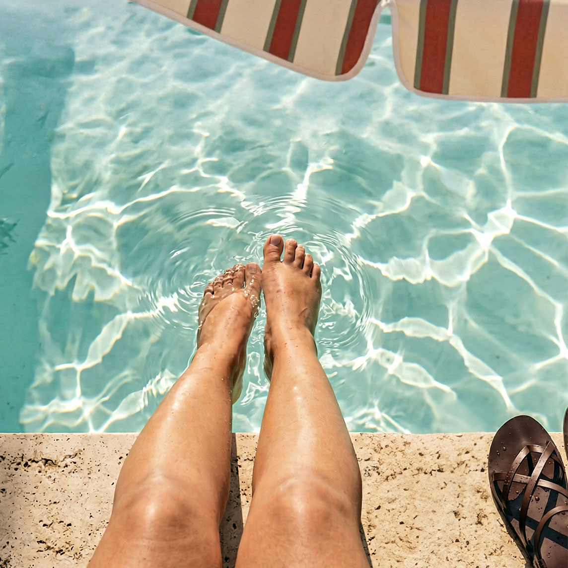 Braune Sandalen liegen am Poolrand, während Füße das kühle Wasser genießen.