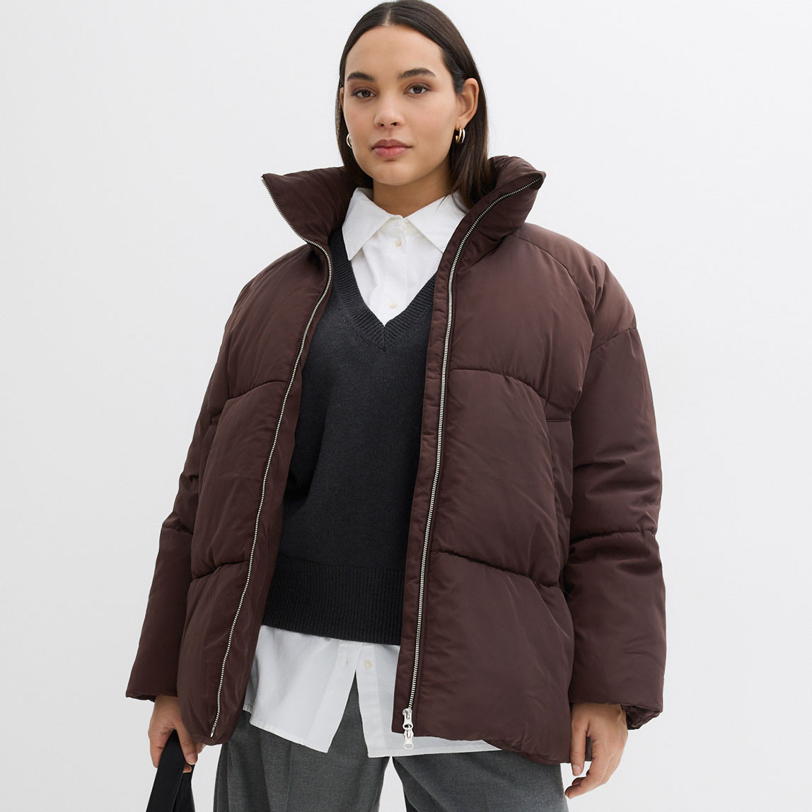 Braune Winterjacke mit hohem Kragen, darunter dunkler Pullover und weißes Hemd.