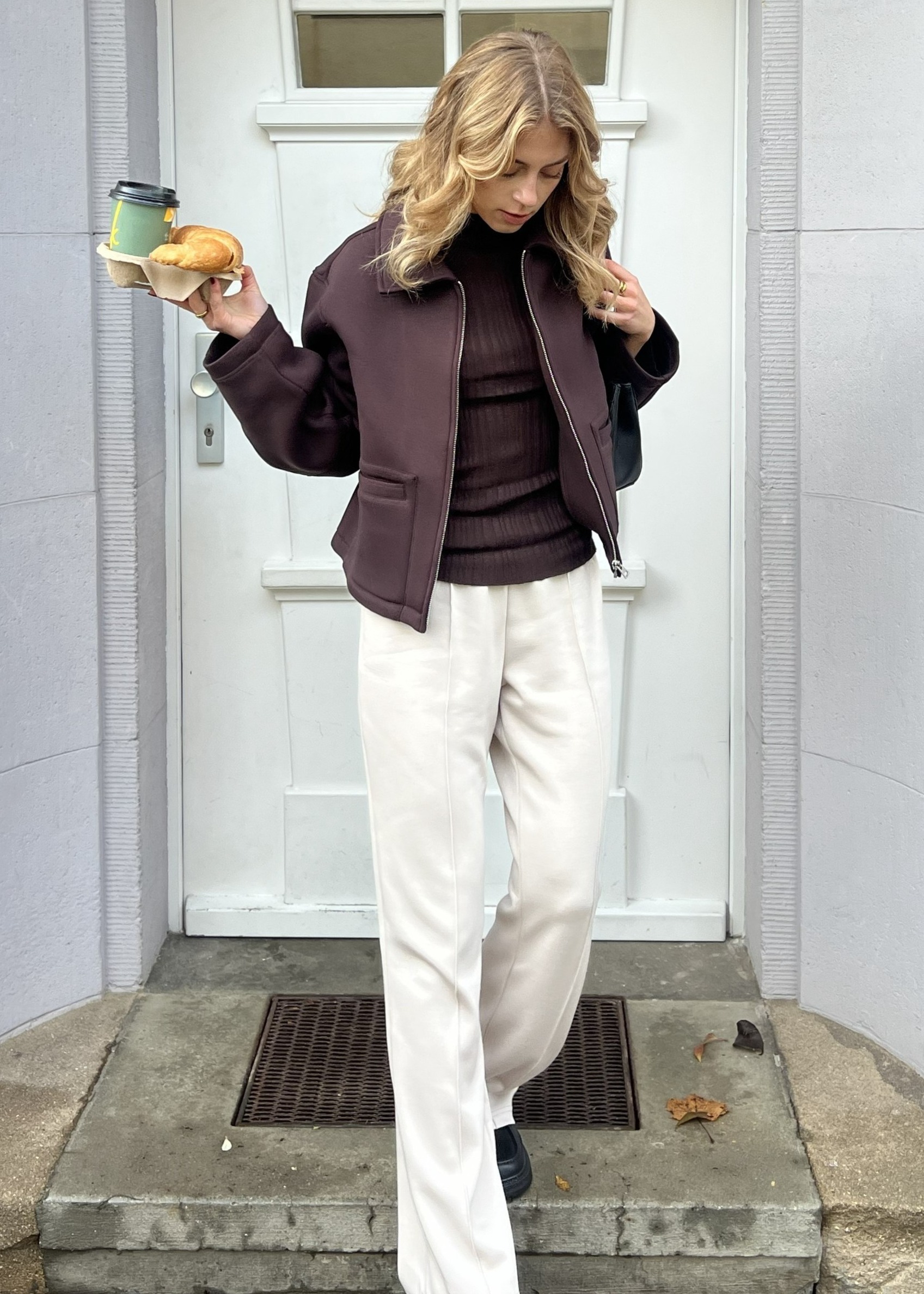 Braune Jacke über dunklem Pullover, kombiniert mit heller, weiter Hose und schwarzen Schuhen.