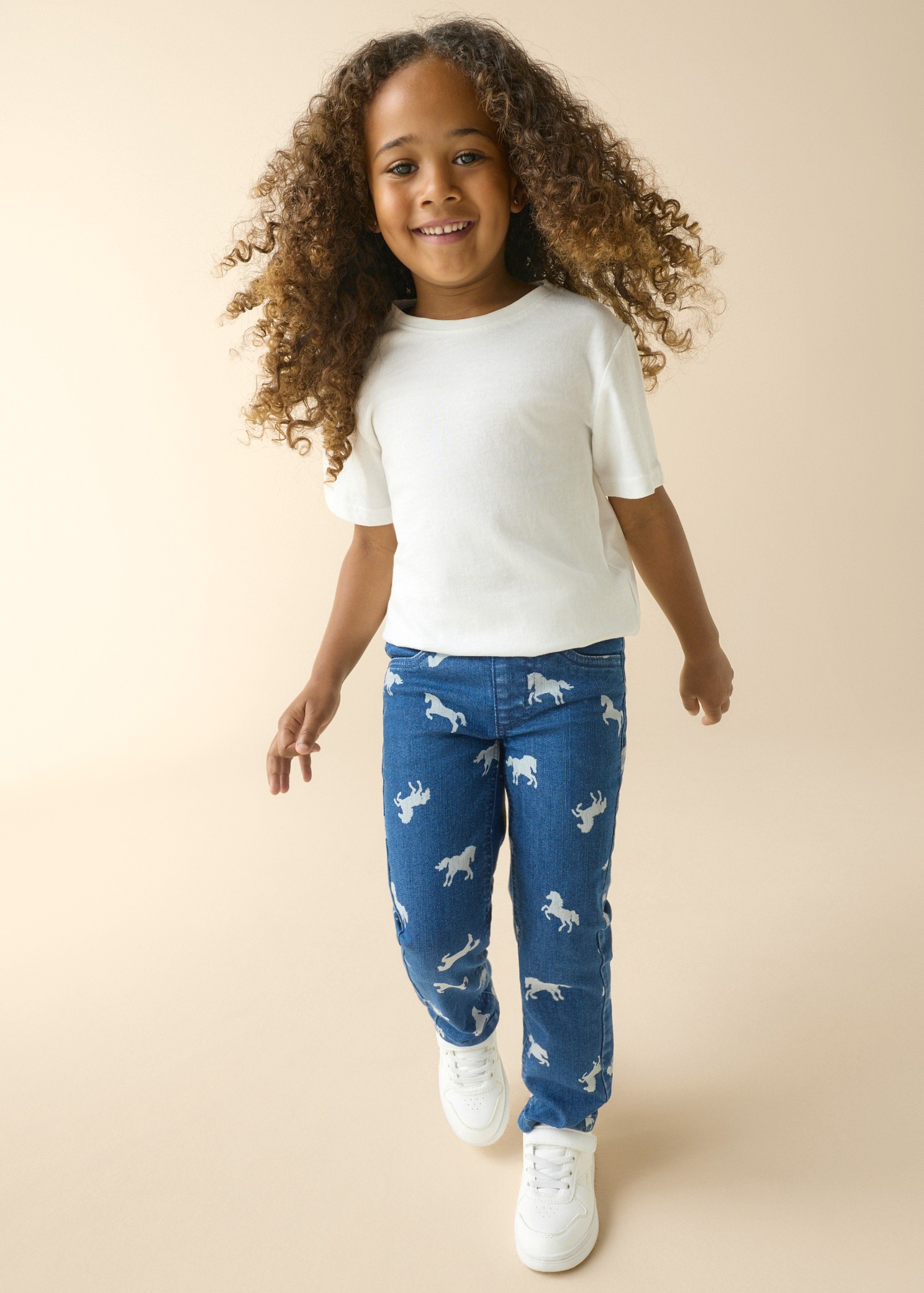 Das Mädchen trägt ein weißes T-Shirt, blaue Jeans mit Pferdemuster und weiße Sneaker.