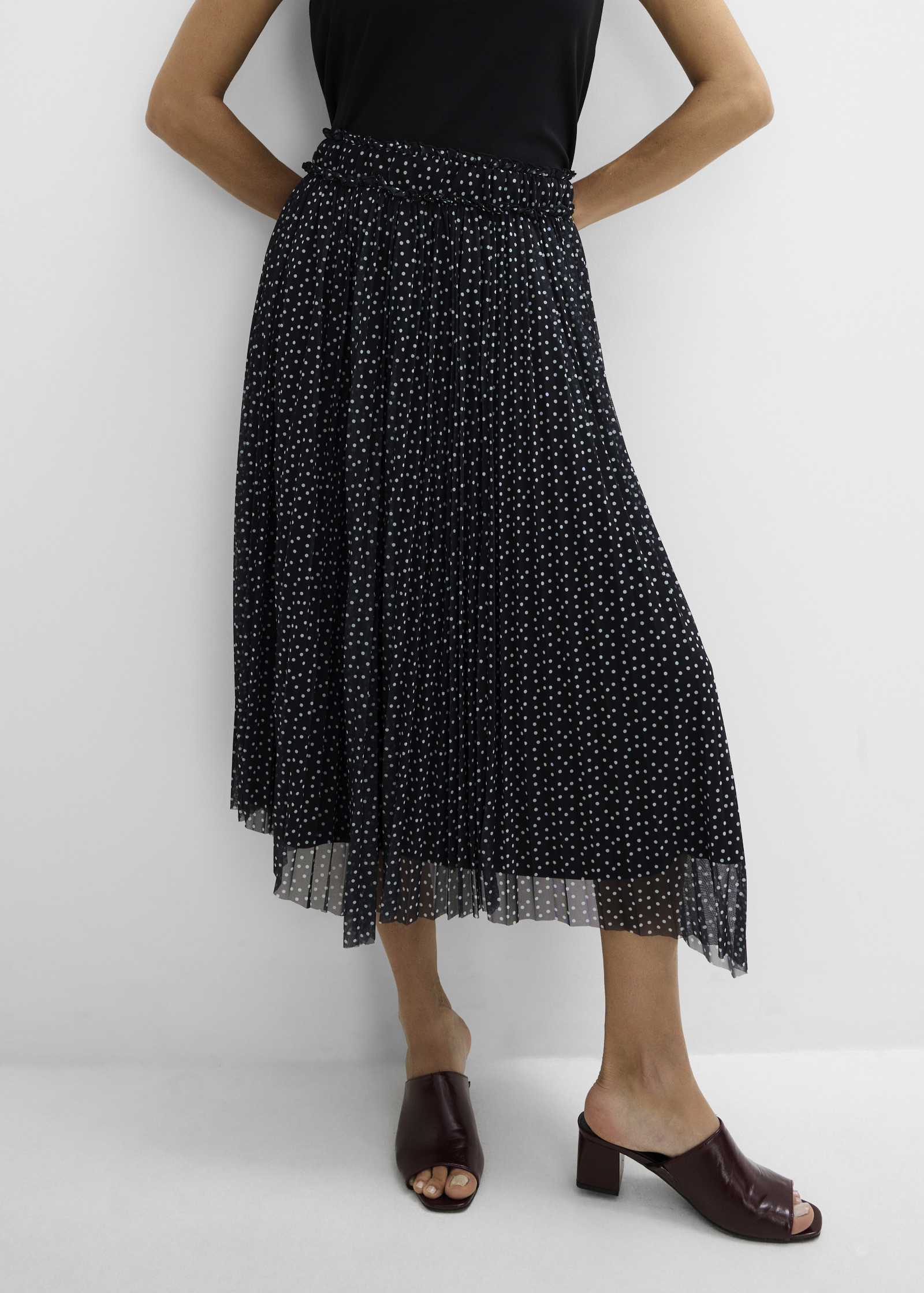 Schlichtes schwarzes Top, gepunkteter Plisseerock und elegante dunkelbraune Pantoletten.