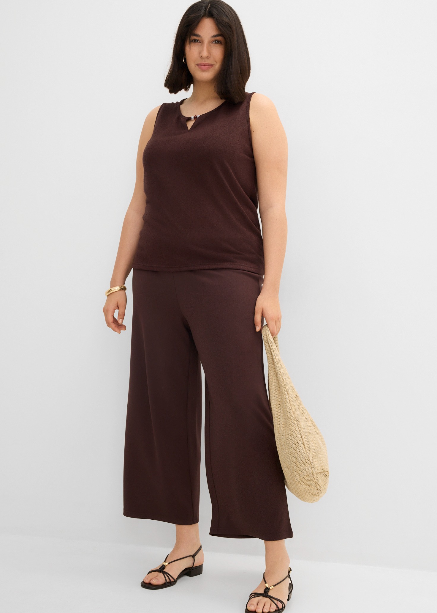 Braunes ärmelloses Top, weite Hose mit schwarzen Sandalen und Korbtasche.