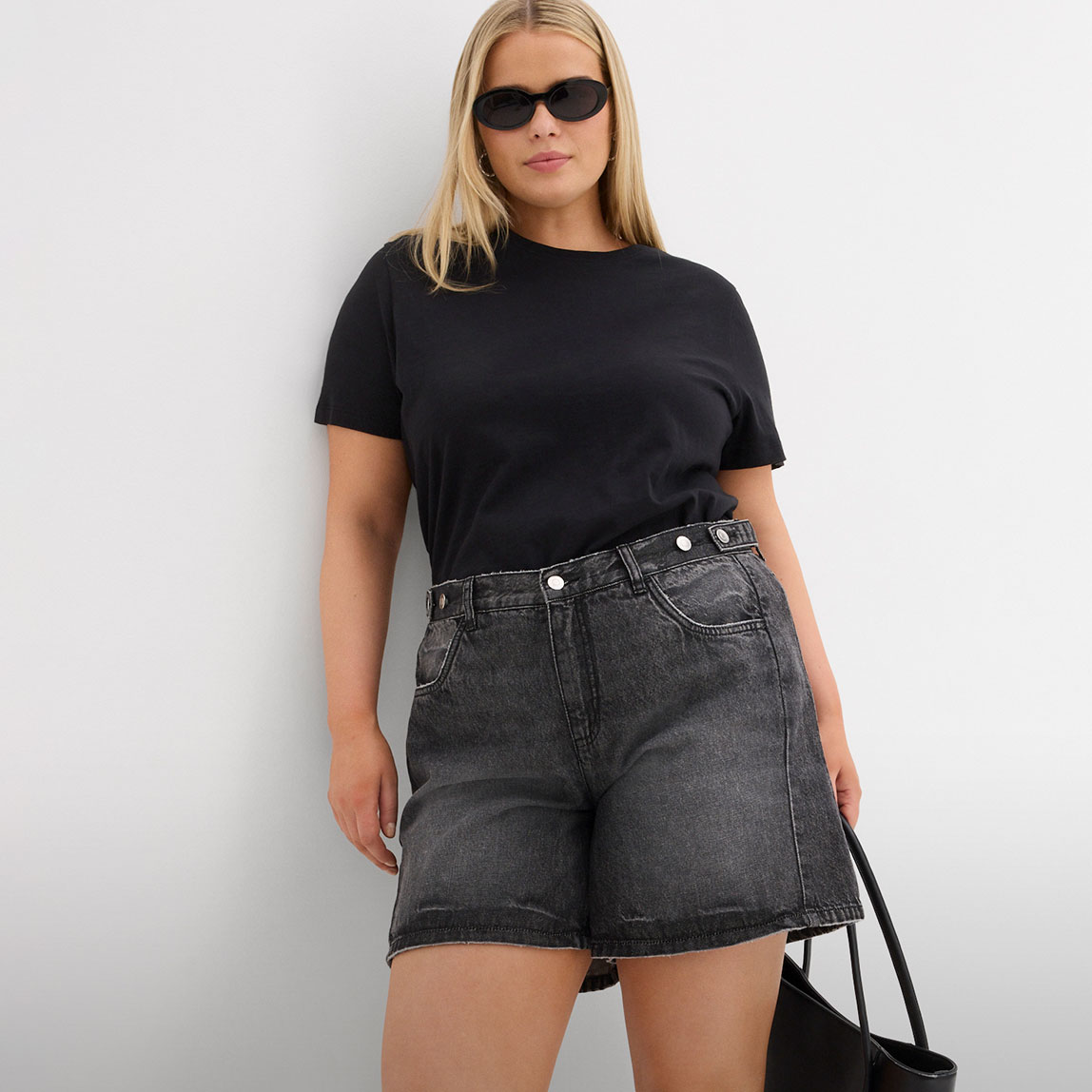 Schwarzes T-Shirt, weite graue Denim-Shorts, Sonnenbrille und schwarze Tasche.