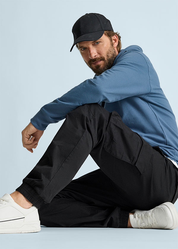 Er trägt eine schwarze Kappe, blauen Hoodie, schwarze Jogger und weiße Sneaker.