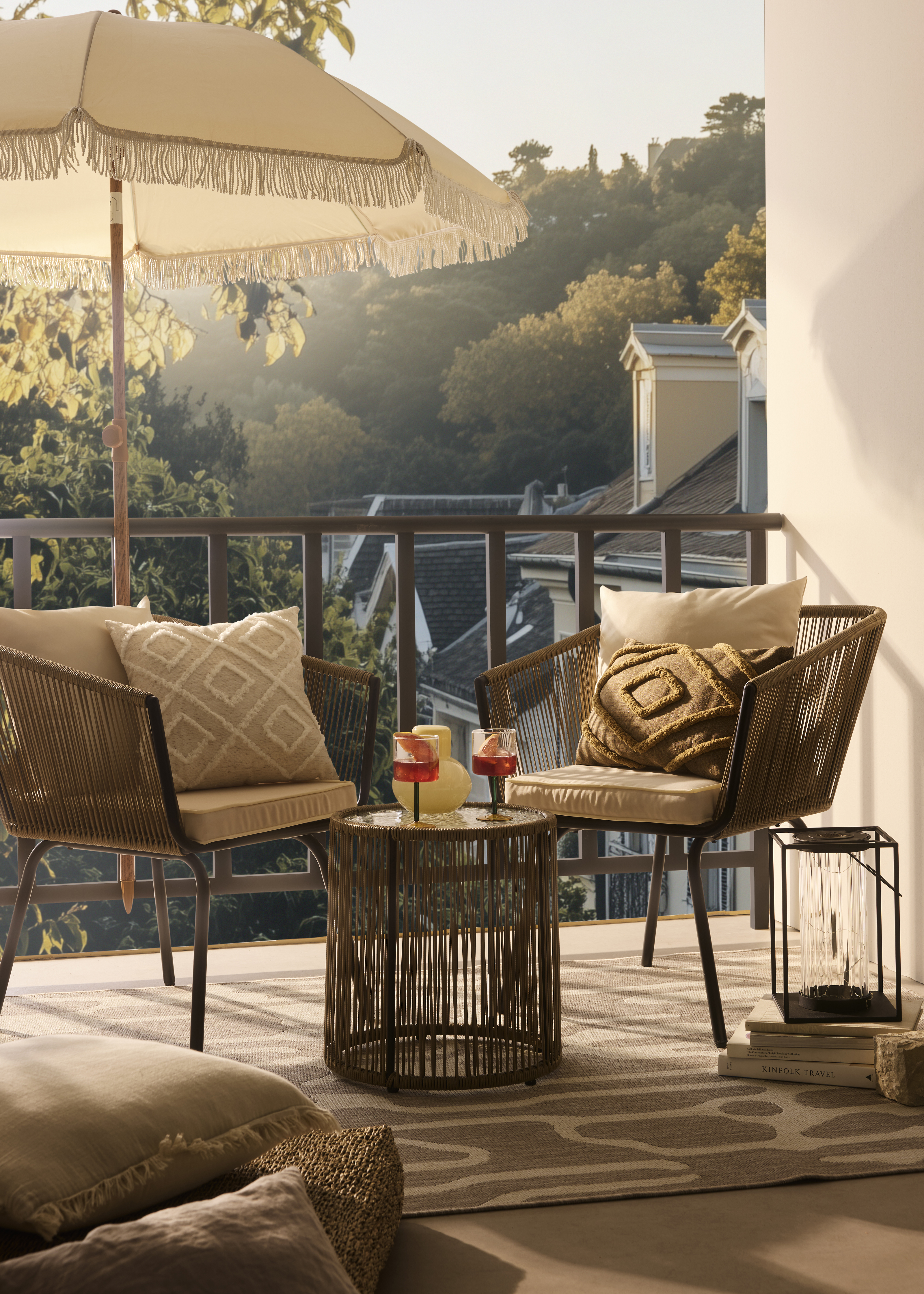 Gemütliche Rattanstühle mit weichen Kissen, beige Sonnenschirm und dekorative Laternen auf stilvoller Terrasse.