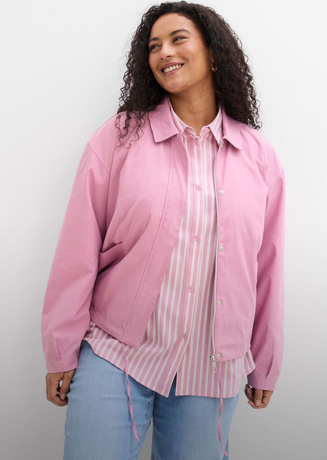 Sie trägt eine rosa Jacke, ein gestreiftes Hemd und helle Jeans.