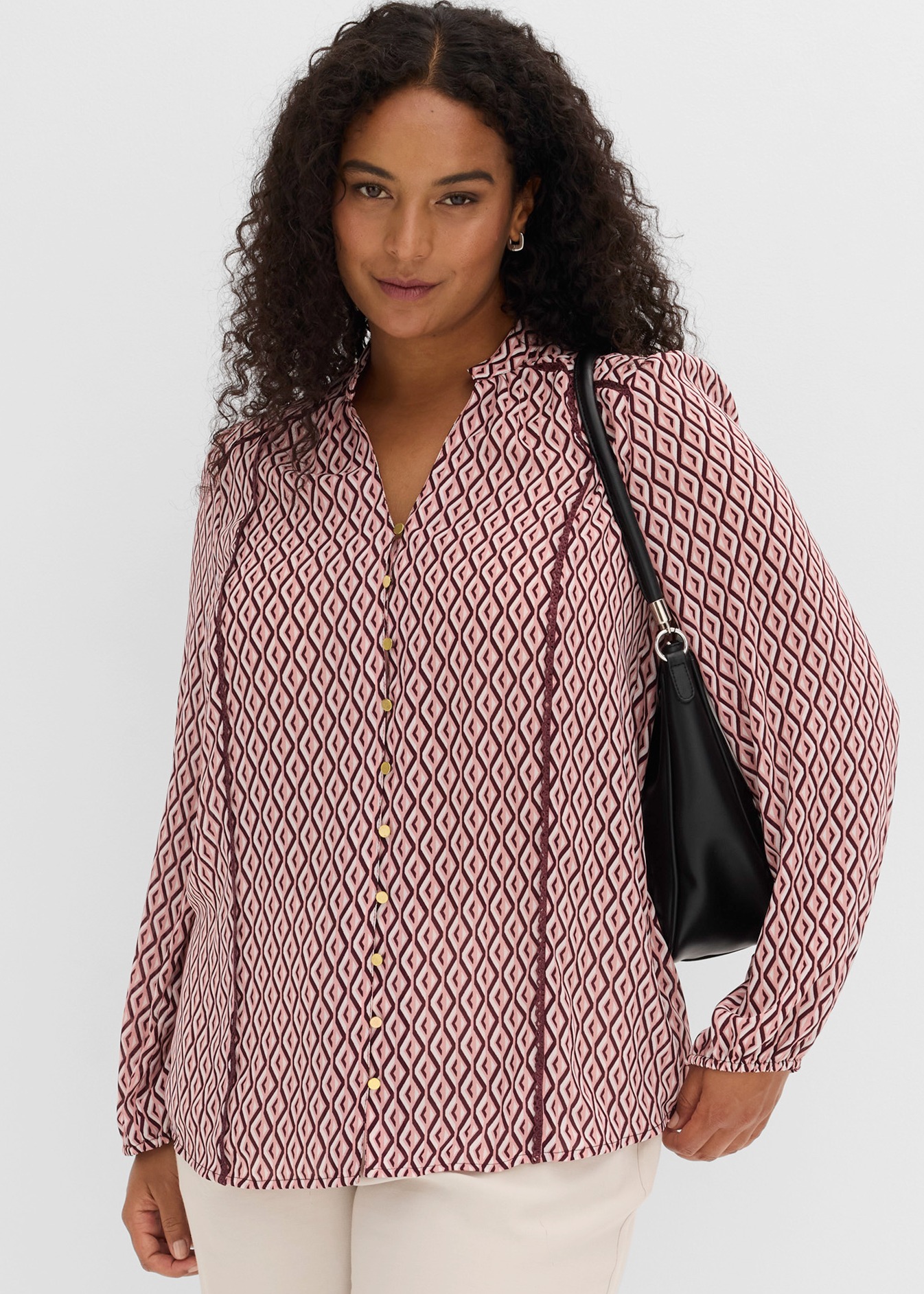 Gemusterte Bluse mit goldenen Knöpfen, helle Hose und schwarze Handtasche.