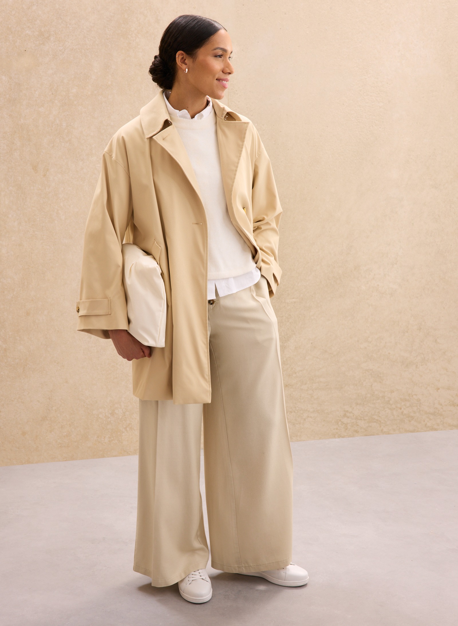 Eleganter beigefarbener Mantel über weißem Pullover, kombiniert mit weiten Hosen und bequemen Sneakern.