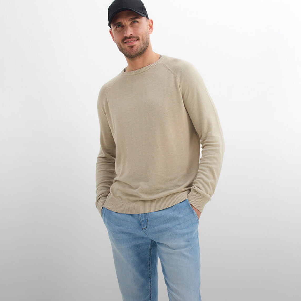 Mann trägt beigen Pullover, blaue Jeans und schwarze Baseballkappe.