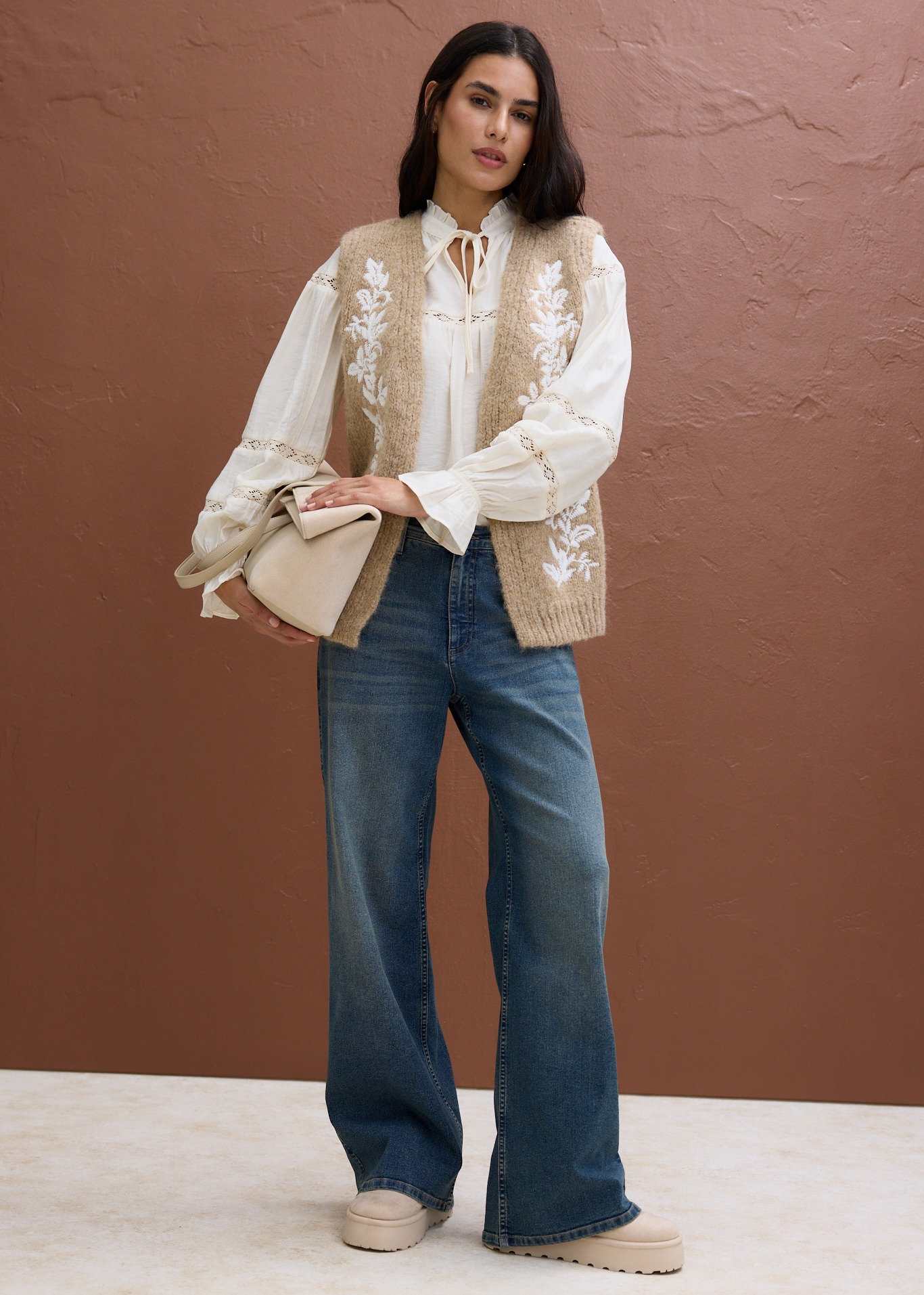 Weiße Bluse mit Spitze, dazu eine beige Strickweste mit floraler Stickerei und Jeans.