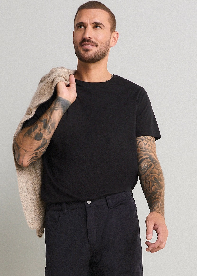 Schwarzes T-Shirt, dunkle Hose und beigefarbener Strickpullover lässig über der Schulter.