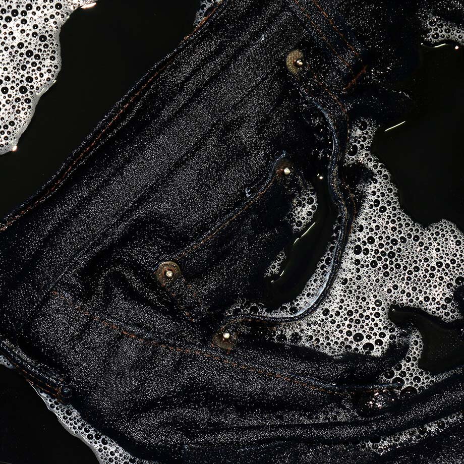 Schwarze Jeans im Wasser, glänzend und nass. Knöpfe und Reißverschluss sichtbar. Schaum umgibt sie.