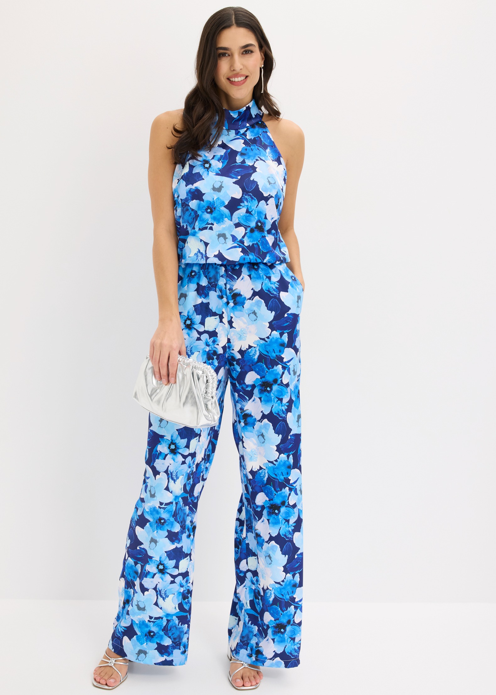 Blauer Overall mit lebhaftem Blumenmuster, elegantem Neckholder und silberner Clutch.