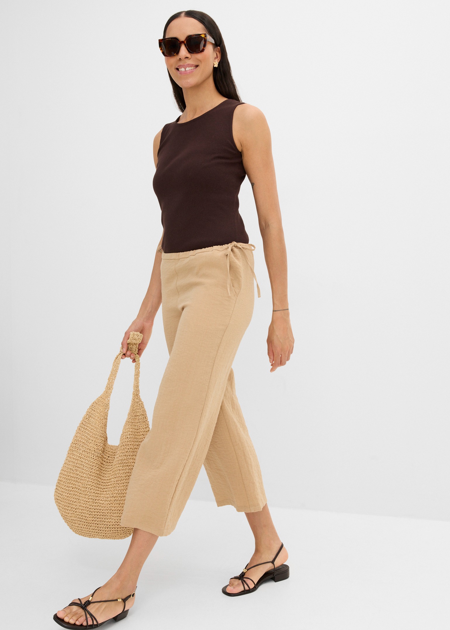 Elegantes braunes Top, weite beigefarbene Hose, schwarze Riemchensandalen und geflochtene Korbtasche.