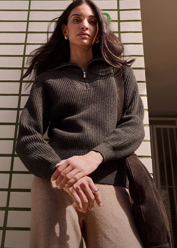 Dunkler Rippstrickpullover mit Reißverschluss, helle Hose und braune Wildledertasche.
