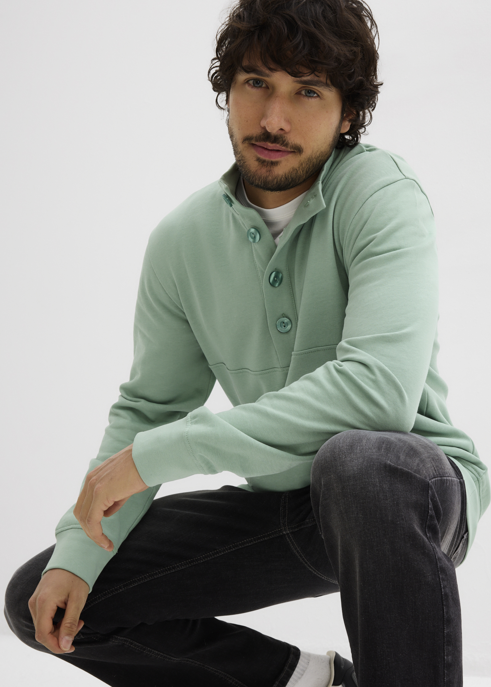 Mintgrünes Sweatshirt mit Knopfleiste, weißes Shirt darunter und dunkle Jeans.