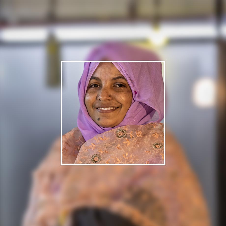 Ein farbenfroher Hijab in Lila schmückt den Kopf. Darunter schimmert ein zartes, gemustertes Oberteil.