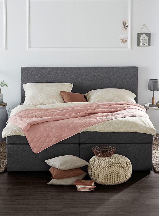 Elegantes Schlafzimmer mit grauem Polsterbett, rosa Tagesdecke und cremefarbenen Kissen. Gestrickter Pouf ergänzt den Raum.