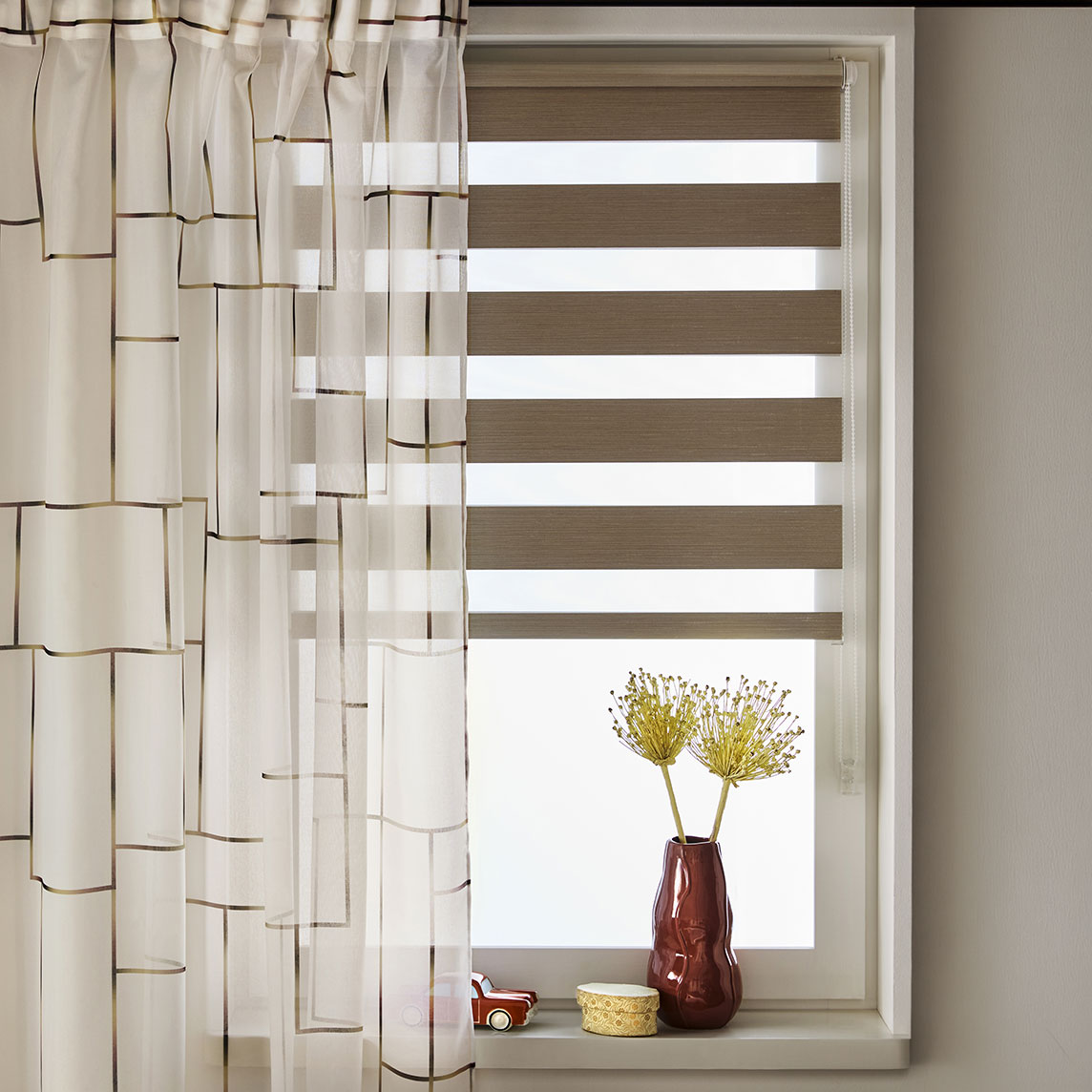 Ein Fenster mit cremefarbener Gardine und einem Plissee, sowie Dekoration auf der Fensterbank.