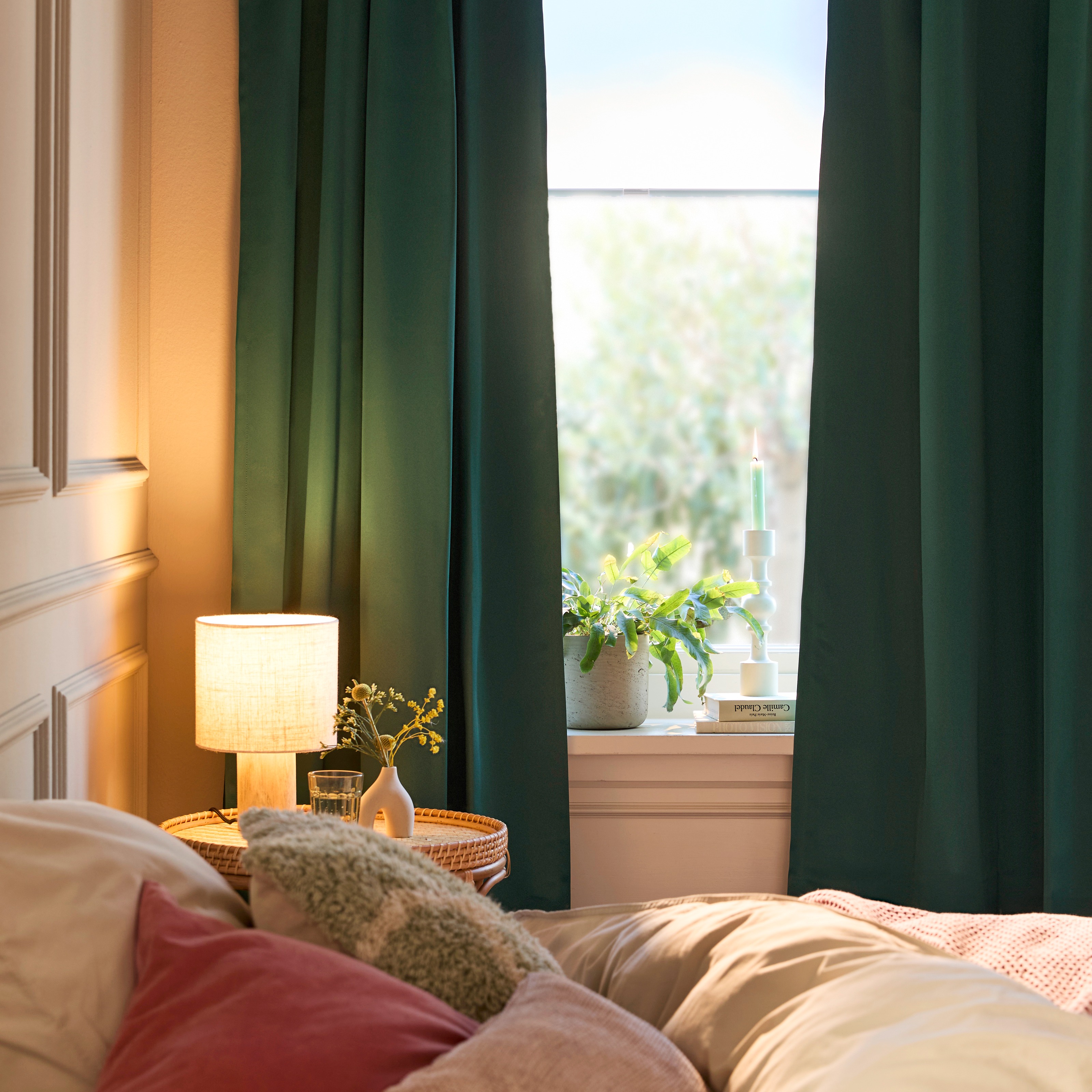 Gemütliches Schlafzimmer mit dunkelgrünen Vorhängen, warmer Beleuchtung und kuscheligen Textilien auf dem Bett.