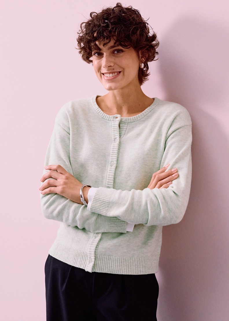 Sie trägt eine hellgrüne Strickjacke zur dunkle Hose.
