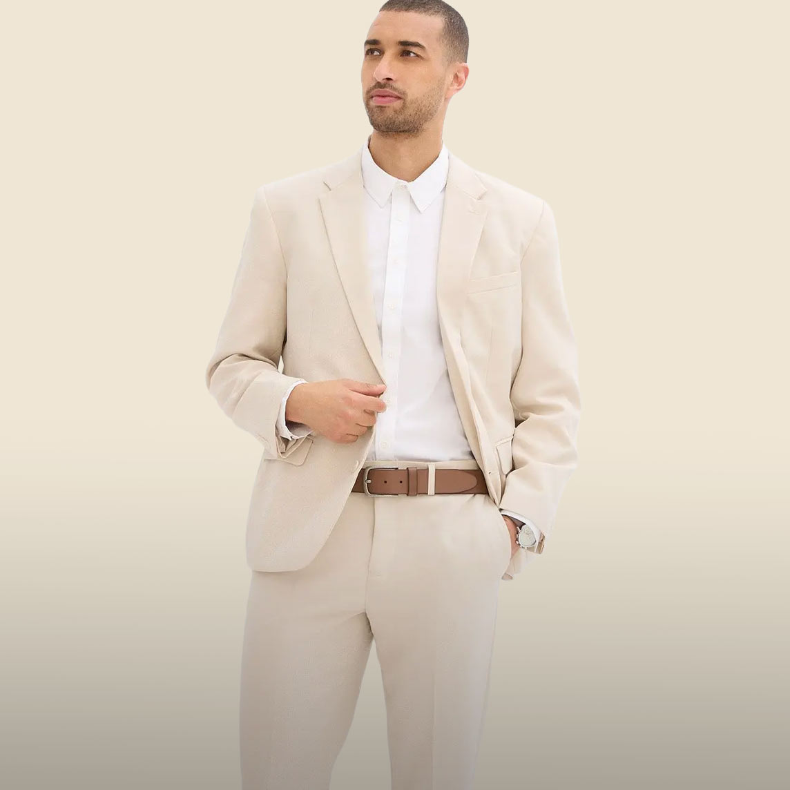 Er trägt einen eleganten beigen Anzug, ein weißes Hemd und einen braunen Gürtel.
